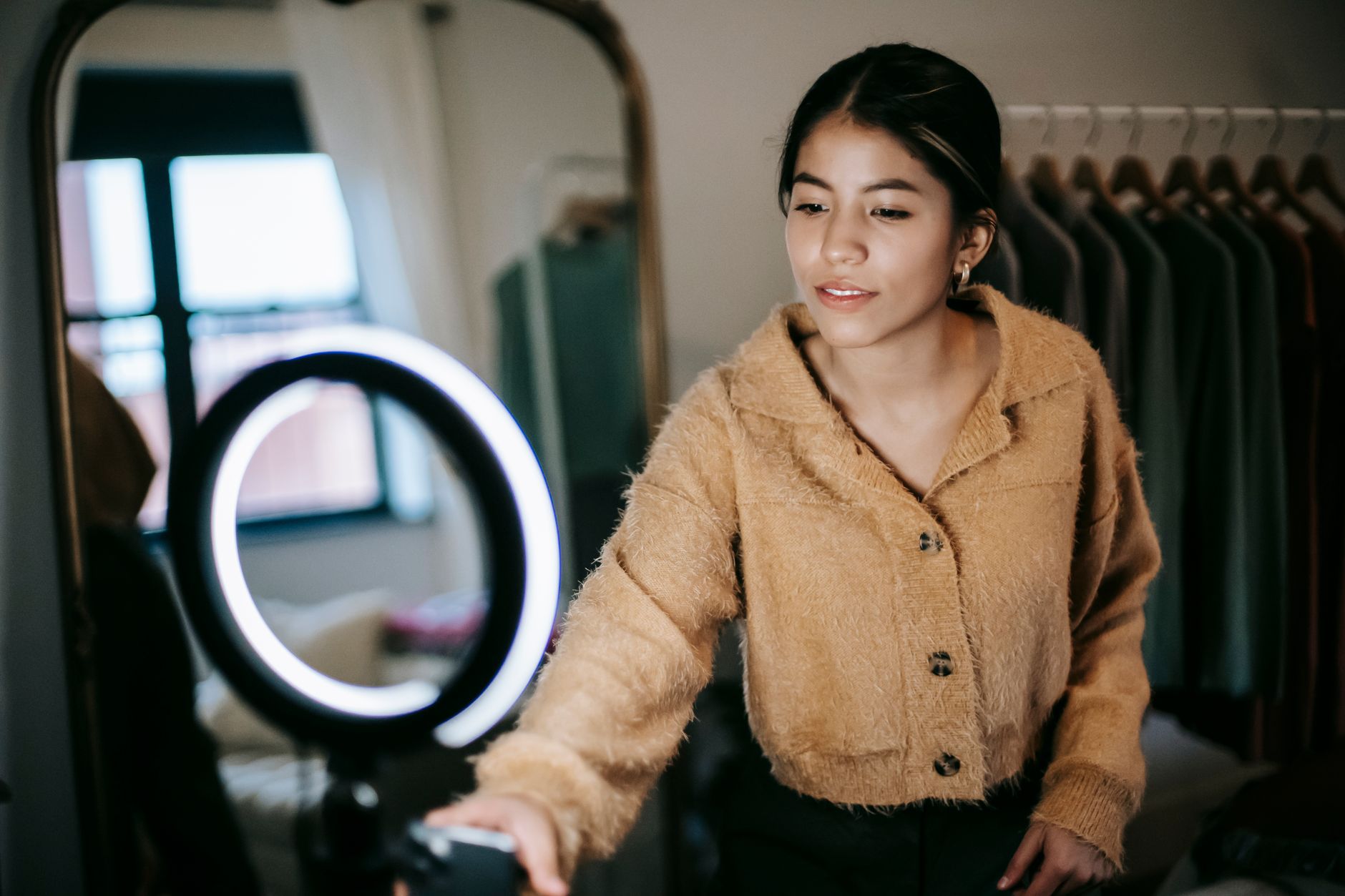 Jeune femme en tenue tendance qui se filme en selfie avec son smartphone sur un anneau lumineux, dans un dressing.